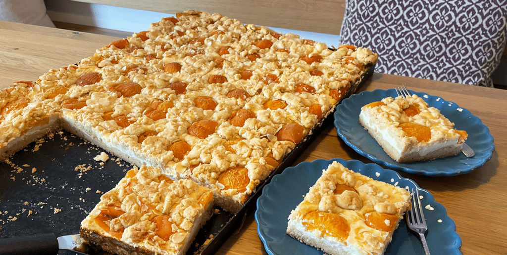 fertig gebackenes Blech mit Aprikosenkuchen. Die Stücke sind angeschnitten und zwei liegen auf jeweils zwei Tellern.