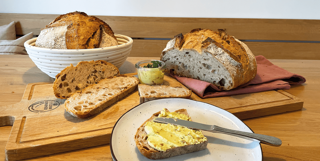 zwei fertig gebackene Dinkel-Landbrote mit aufgeschnitten Scheiben und einer bestrichenen Scheibe im Vordergrund