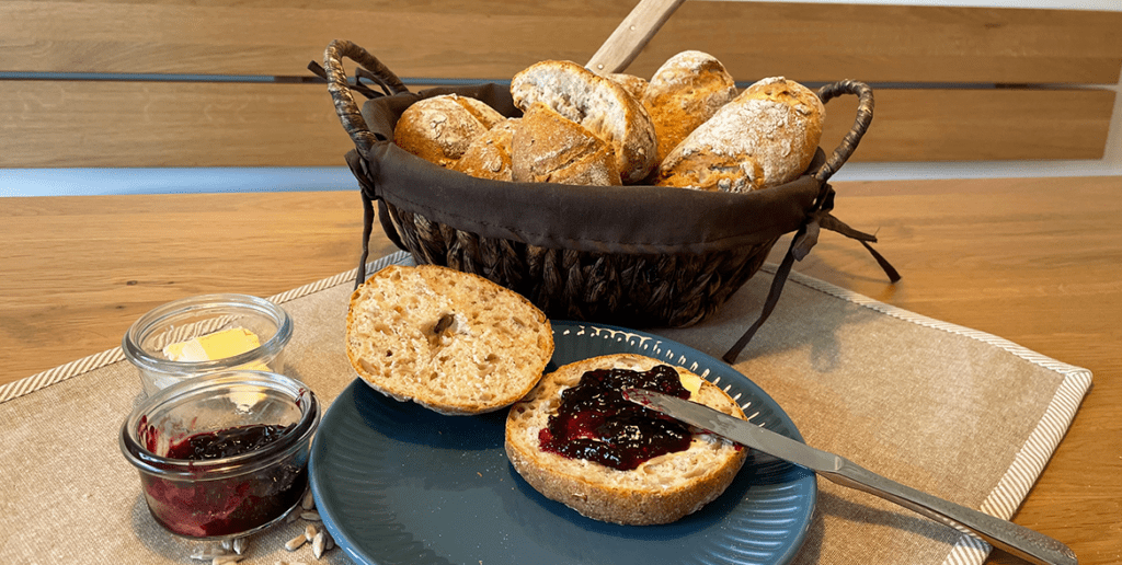 Im Hintergrund steht ein Brötchenkorb mit Sonnenblumenkern-Schrotbrötchen. Im Vordergrund ist ein Brötchen aufgeschnitten und mit Marmelade bestrichen.