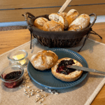 Im Hintergrund steht ein Brötchenkorb mit Sonnenblumenkern-Schrotbrötchen. Im Vordergrund ist ein Brötchen aufgeschnitten und mit Marmelade bestrichen.