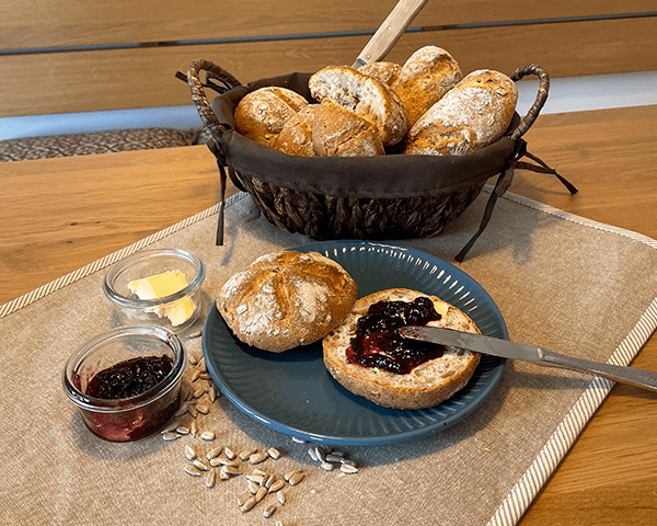 Im Hintergrund steht ein Brötchenkorb mit Sonnenblumenkern-Schrotbrötchen. Im Vordergrund ist ein Brötchen aufgeschnitten und mit Marmelade bestrichen.