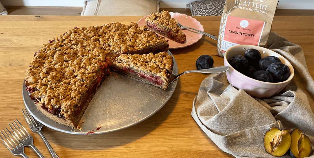 Rezeptvariation aus der Lintertorten Backmischung verfeinert mit Zwetschgen und Preiselbeeren. Auf dem Bild ist der Kuchen angeschnitten und die Verpackung der Backmischung sowie Deko steht auf dem Tisch.