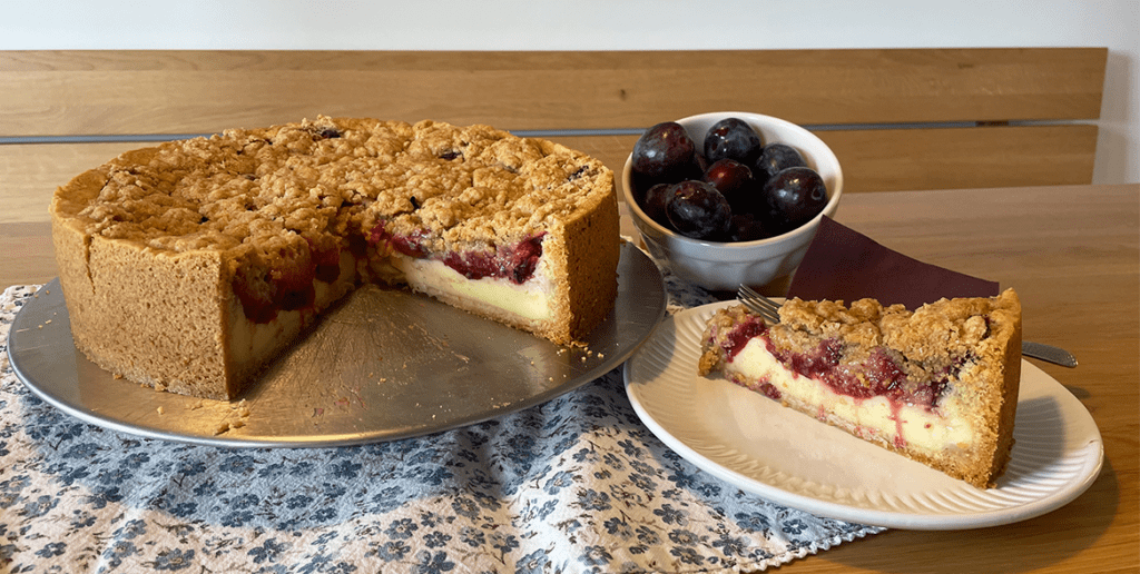 frisch gebackener Zwetschgenkuchen mit Puddingcreme und Zimtstreuseln. Der Kuchen ist angeschnitten und ein Stück steht auf einem Teller daneben.
