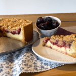 frisch gebackener Zwetschgenkuchen mit Puddingcreme und Zimtstreuseln. Der Kuchen ist angeschnitten und ein Stück steht auf einem Teller daneben.