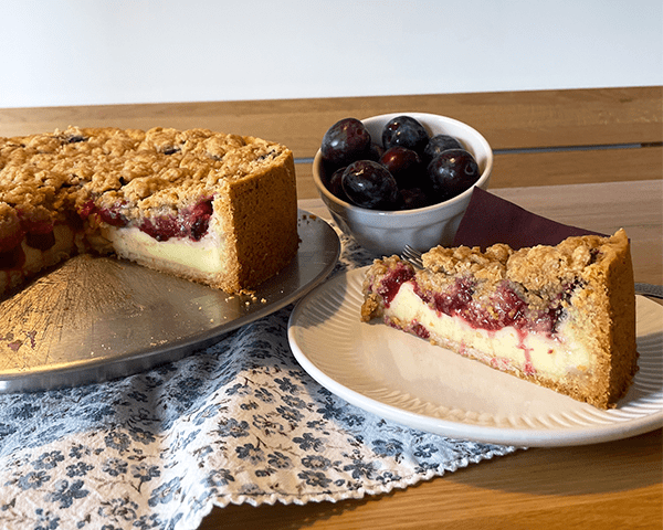 frisch gebackener Zwetschgenkuchen mit Puddingcreme und Zimtstreuseln. Der Kuchen ist angeschnitten und ein Stück steht auf einem Teller daneben.
